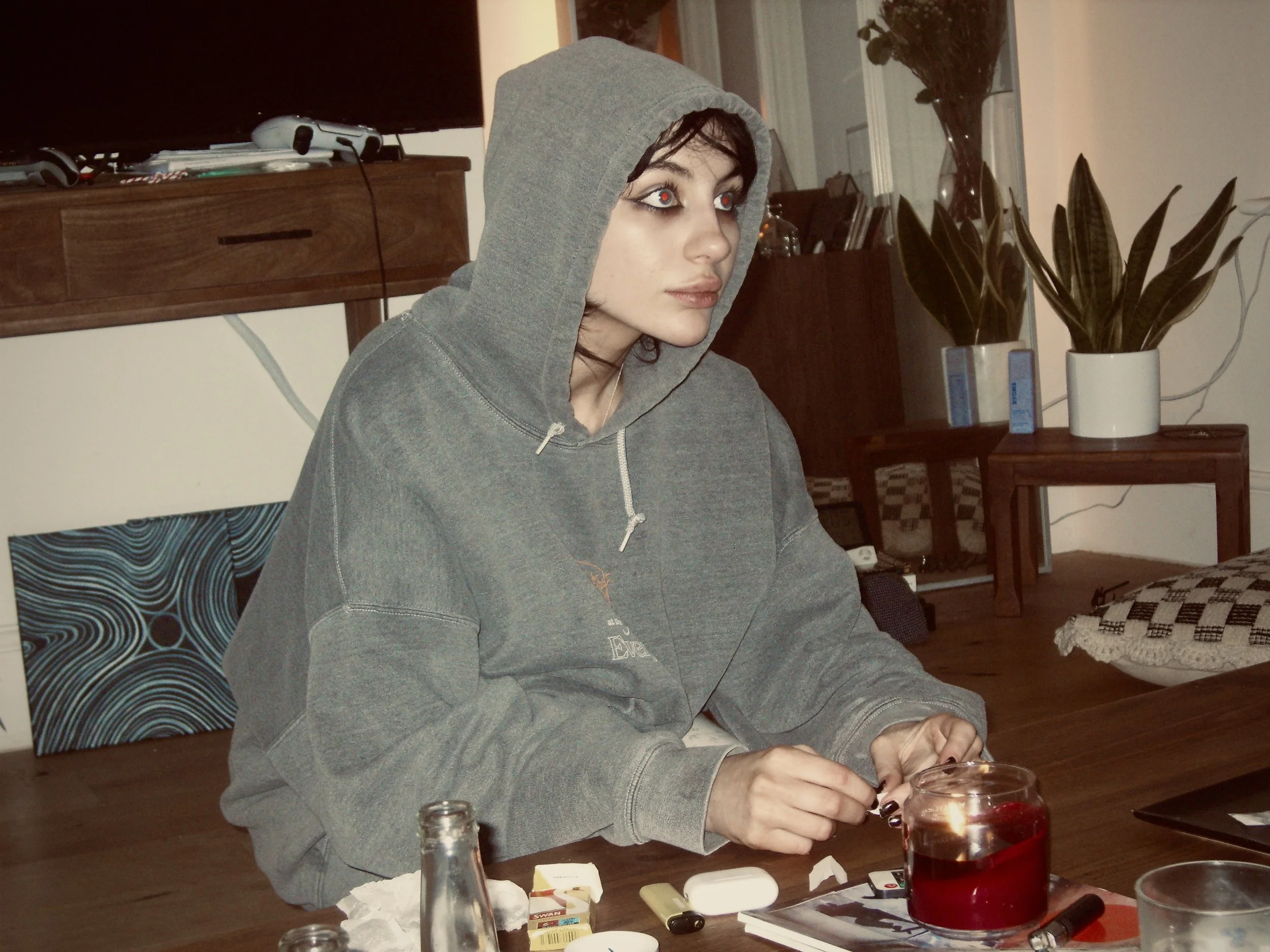 A woman with dark hair wearing a gray hoodie with the hood up, sitting at a wooden table with various objects, including a red lit candle, a bottle, some cards, and other items, in a room with a large potted plant and furniture.