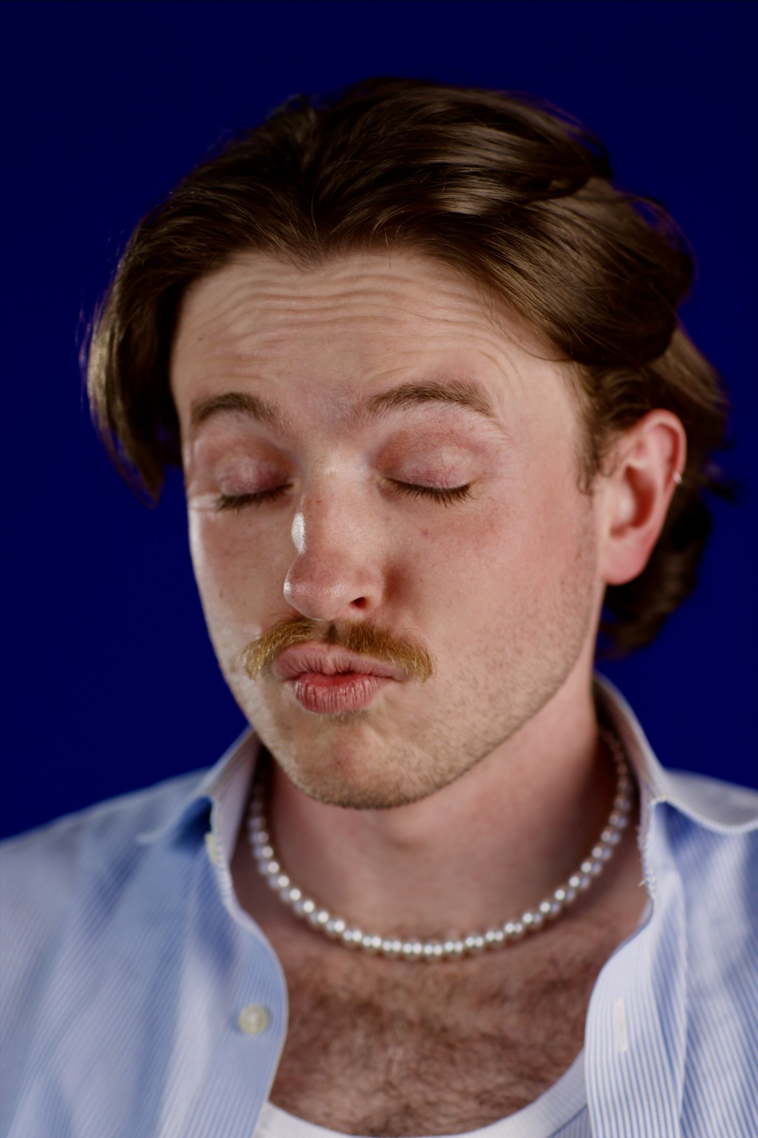 Close-up of a man with closed eyes, light brown hair, a mustache, wearing a pearl necklace and a light blue striped shirt with the top button undone, against a dark blue background.
