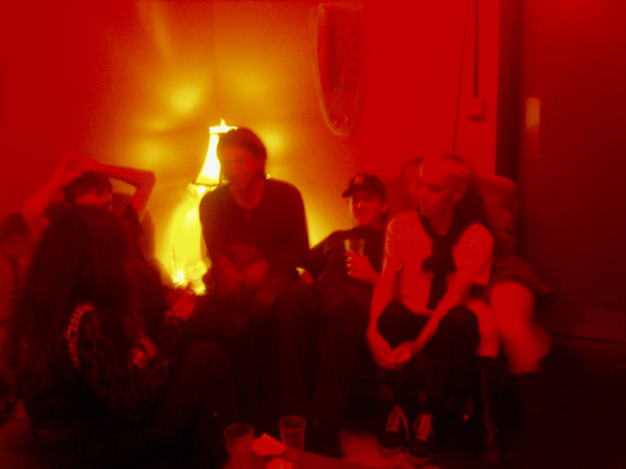 A group of people standing together at a party or nightclub with red lighting, some holding drinks.