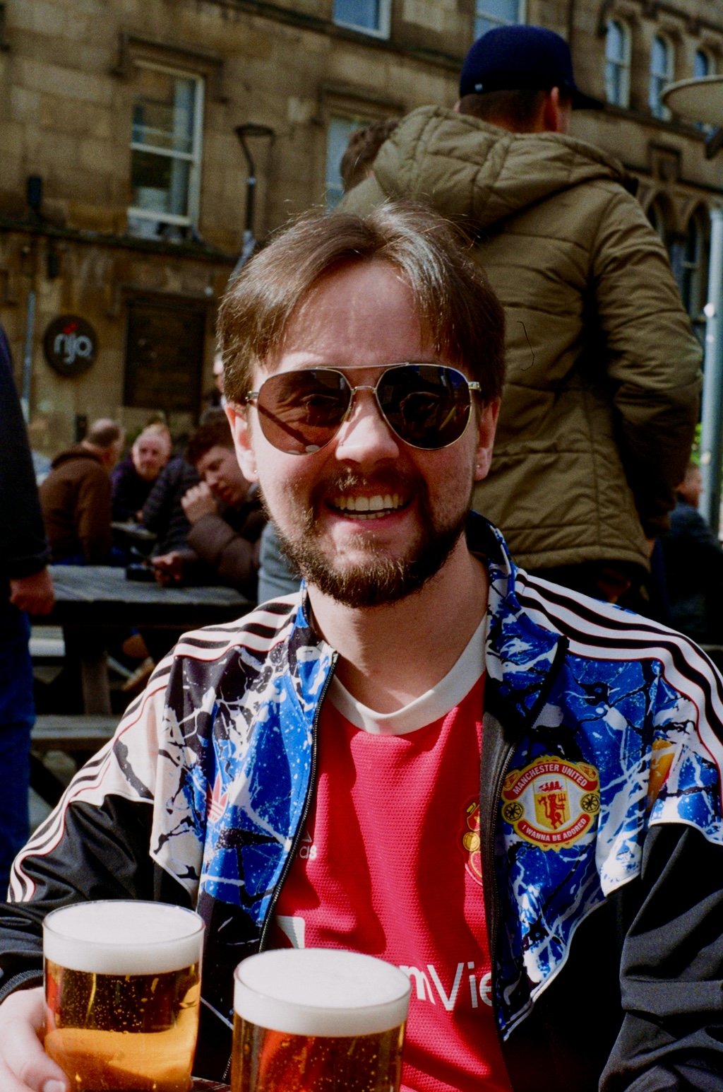 A smiling man wearing sunglasses, a Manchester United football jersey, and a patterned jacket sitting at an outdoor bar or restaurant, with two glasses of beer in front of him, in a lively social setting with other people in the background.