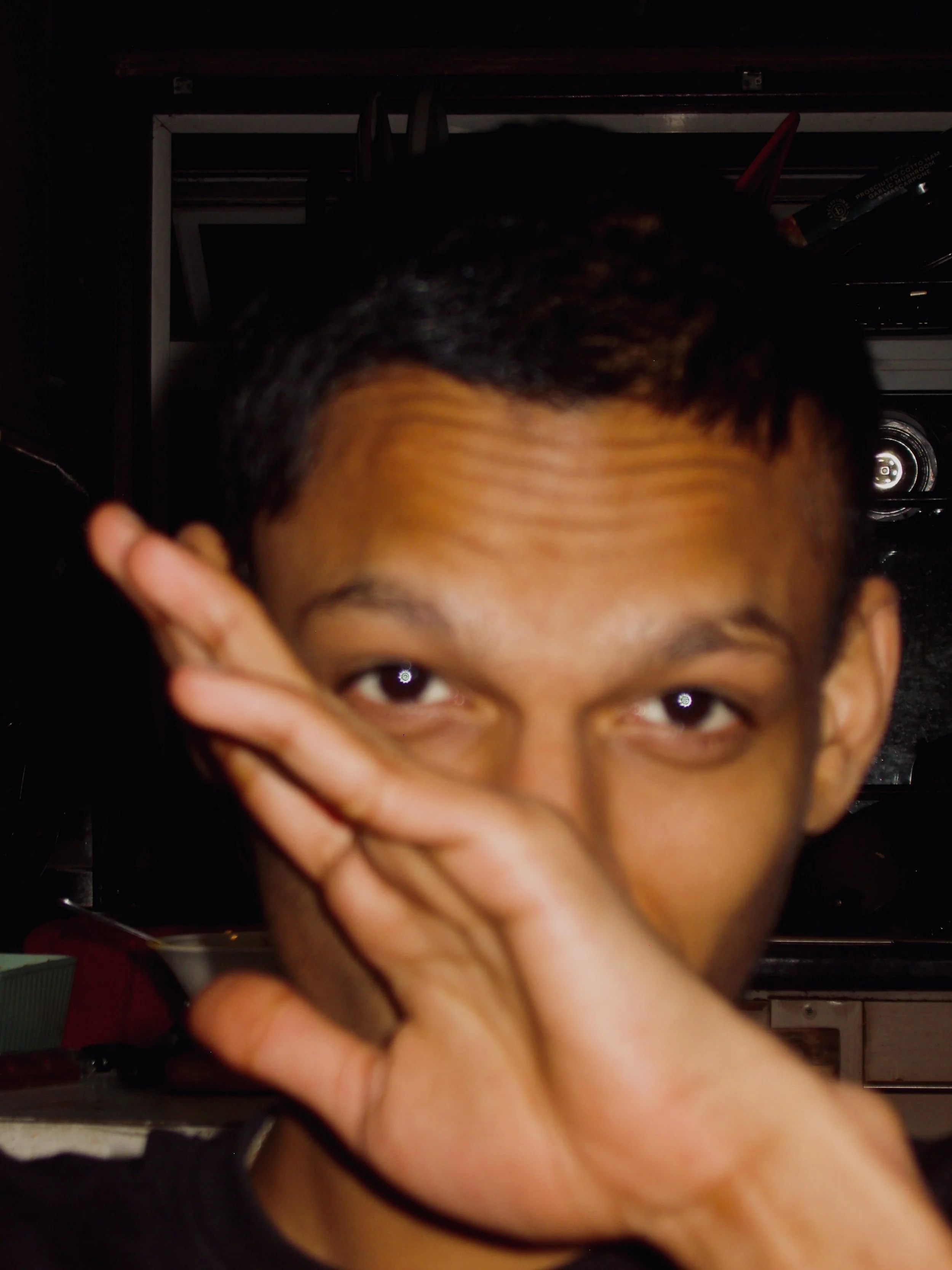 Close-up of a person with short black hair and brown skin, partially obscured by their hand making a peace sign over their face, in a dimly lit room.