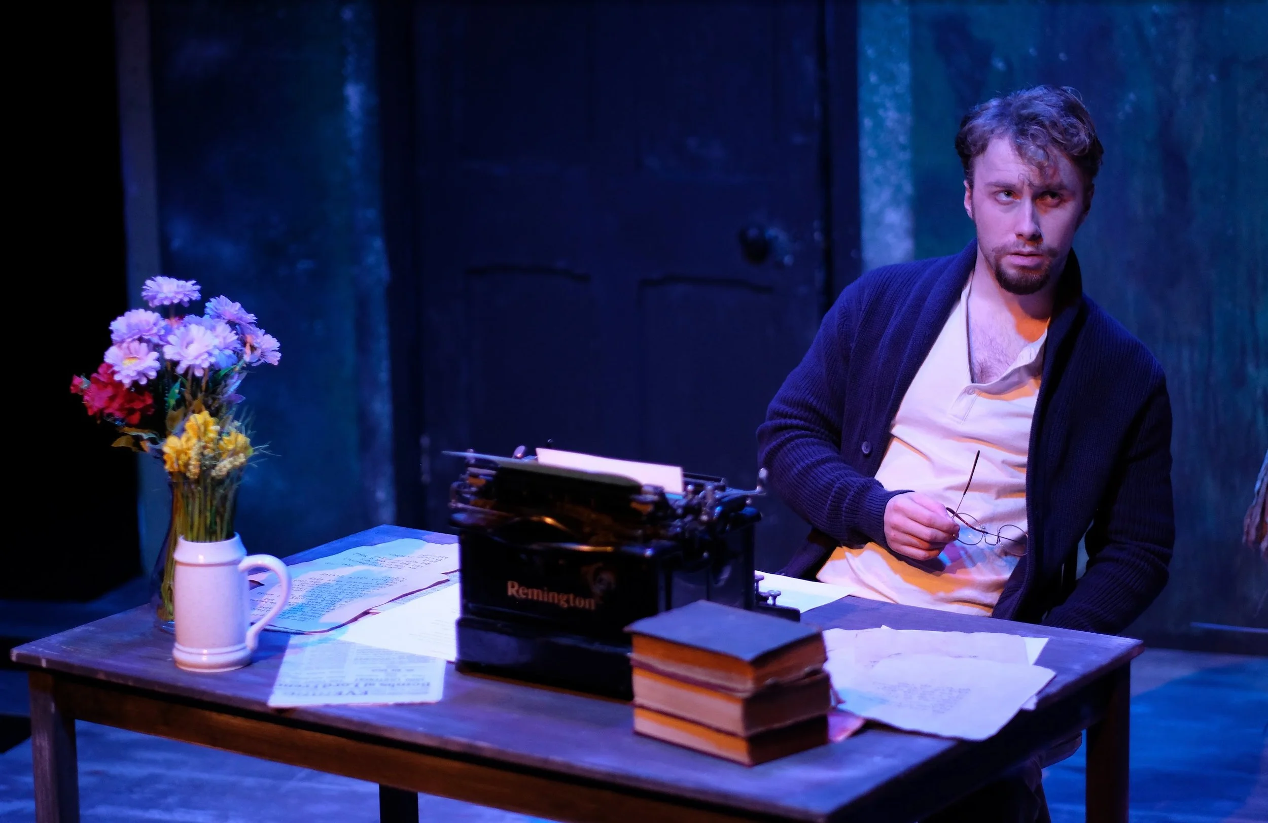 A man sitting at a wooden desk with papers, a stack of books, a typewriter, and a vase of colorful flowers, in a dimly lit room with blue-toned lighting.