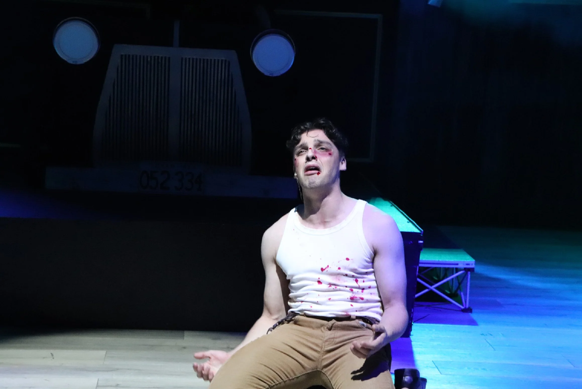 A man with disheveled hair and a distressed expression, wearing a white tank top with blood stains, sitting on a stage with a dark background and stage equipment behind him.
