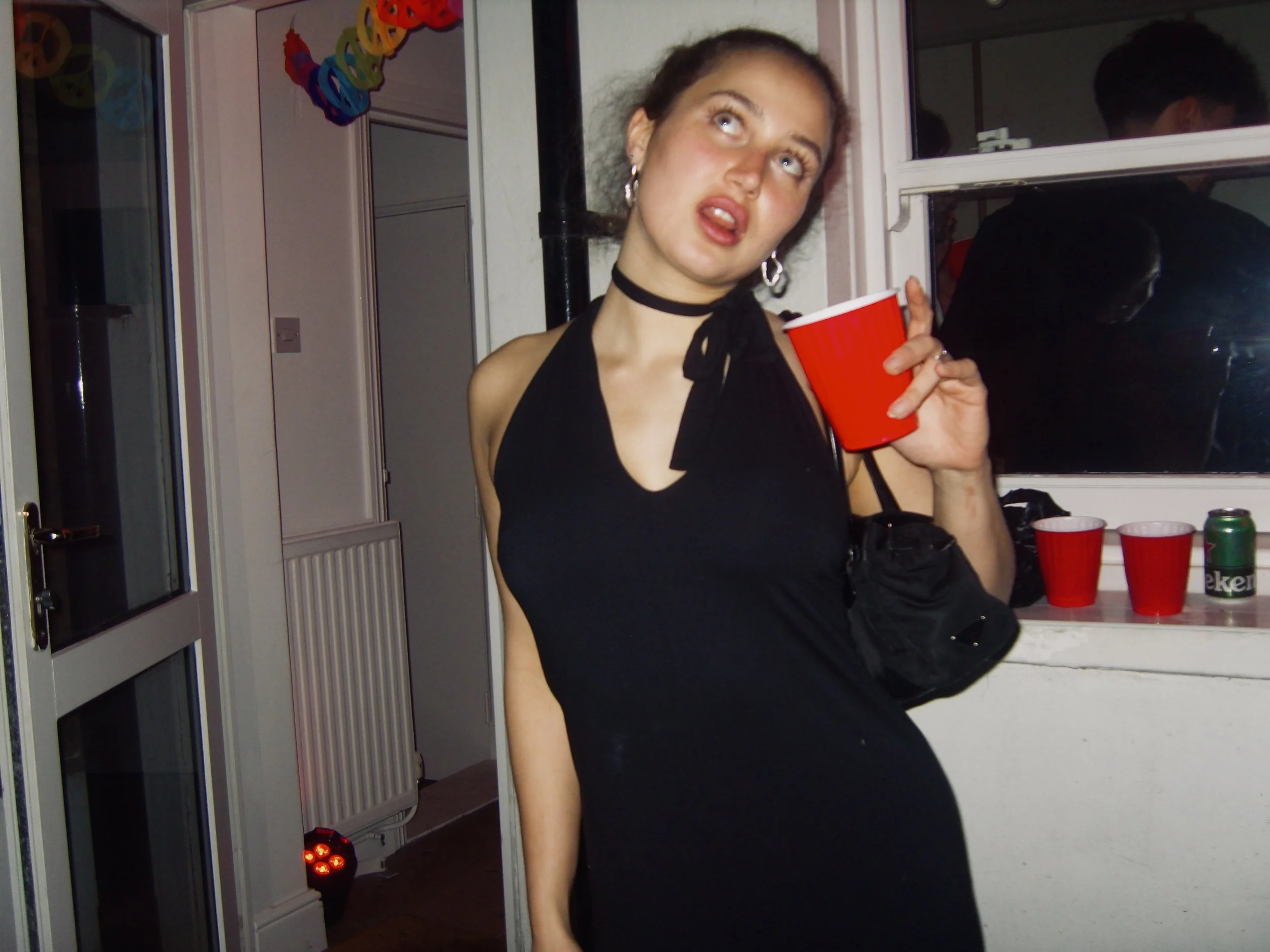 A young woman with light-colored eyes and curly hair holds a red plastic cup, calendar in the background, wearing a black sleeveless top with a bow at the neckline, and hoop earrings, at a party or social gathering.