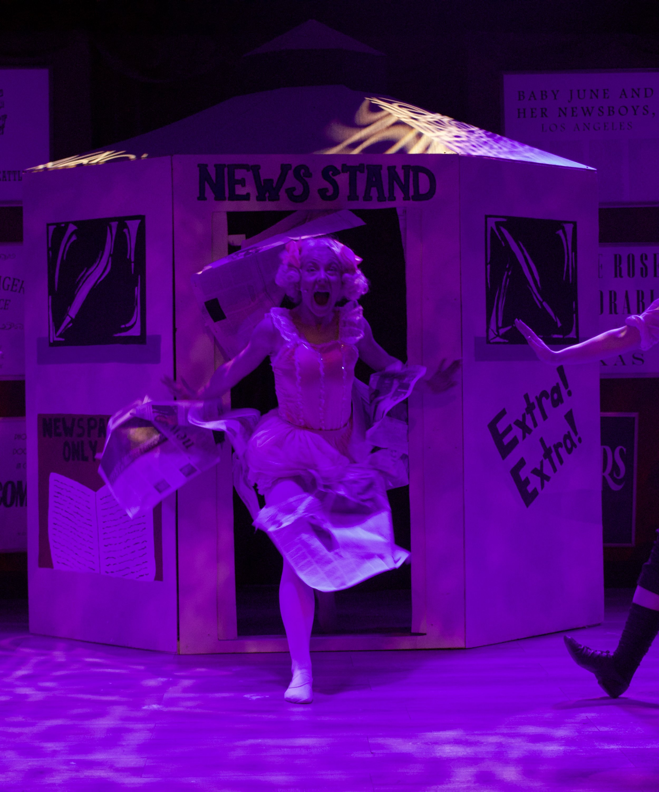 A performer dressed as a newspaper person in a theatrical costume with a wig, performing a dance on stage with a set resembling a newspaper stand, illuminated by purple stage lighting.