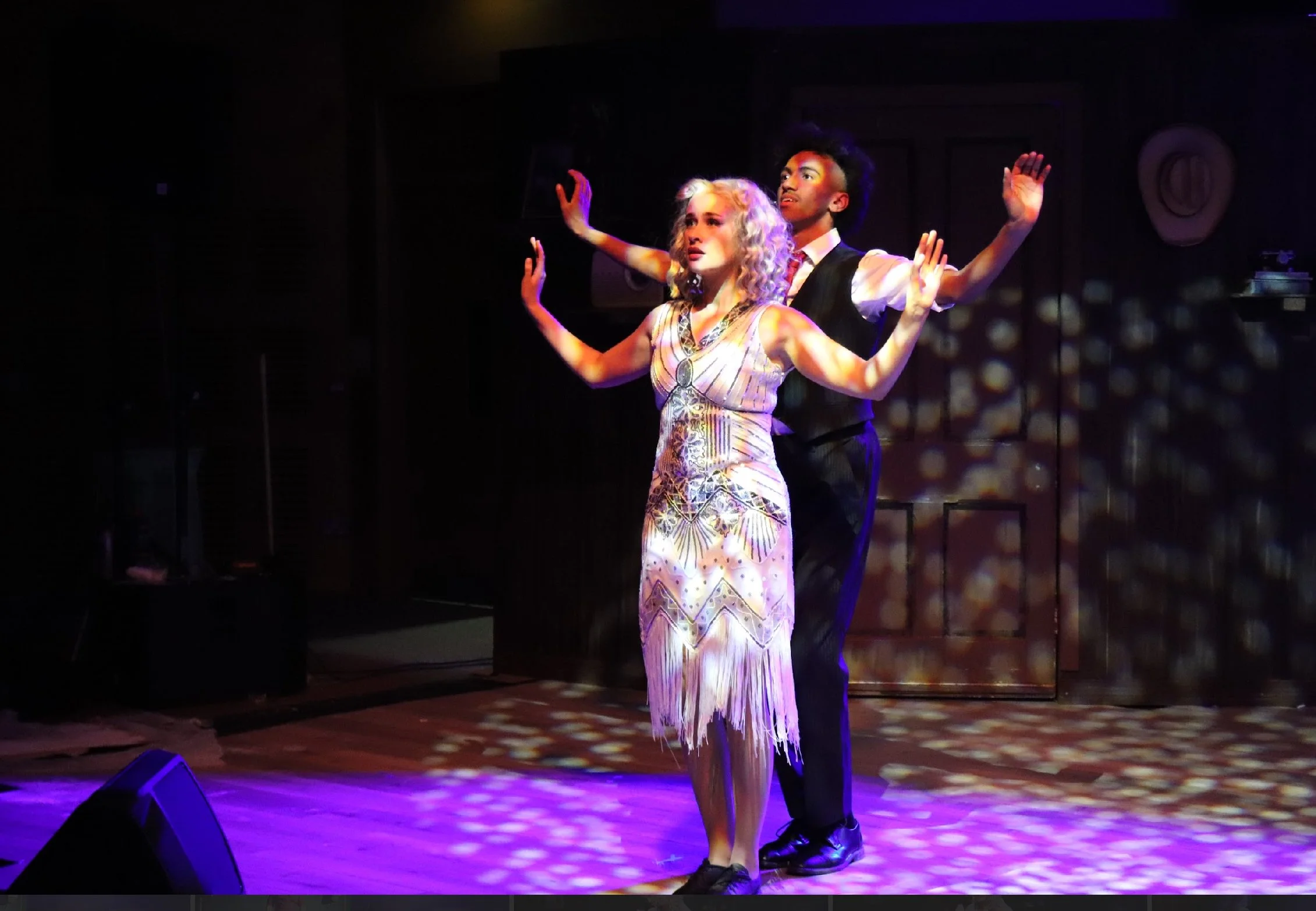 Two performers on stage, a woman in a fringed dress with curly blonde hair and a man in a shirt and vest with an afro hairstyle, dancing under colorful stage lights.