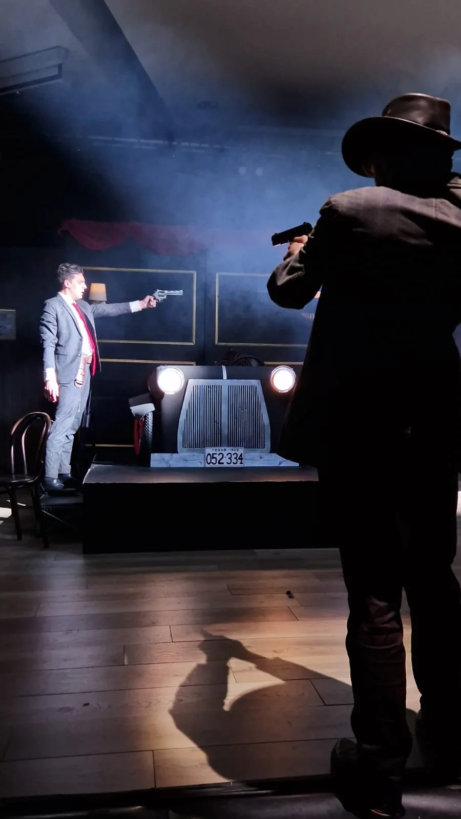 A theatrical scene showing two actors, one in a suit and the other in cowboy attire, aiming guns at each other on a stage with a vintage car, dim lighting, and a smoky atmosphere.