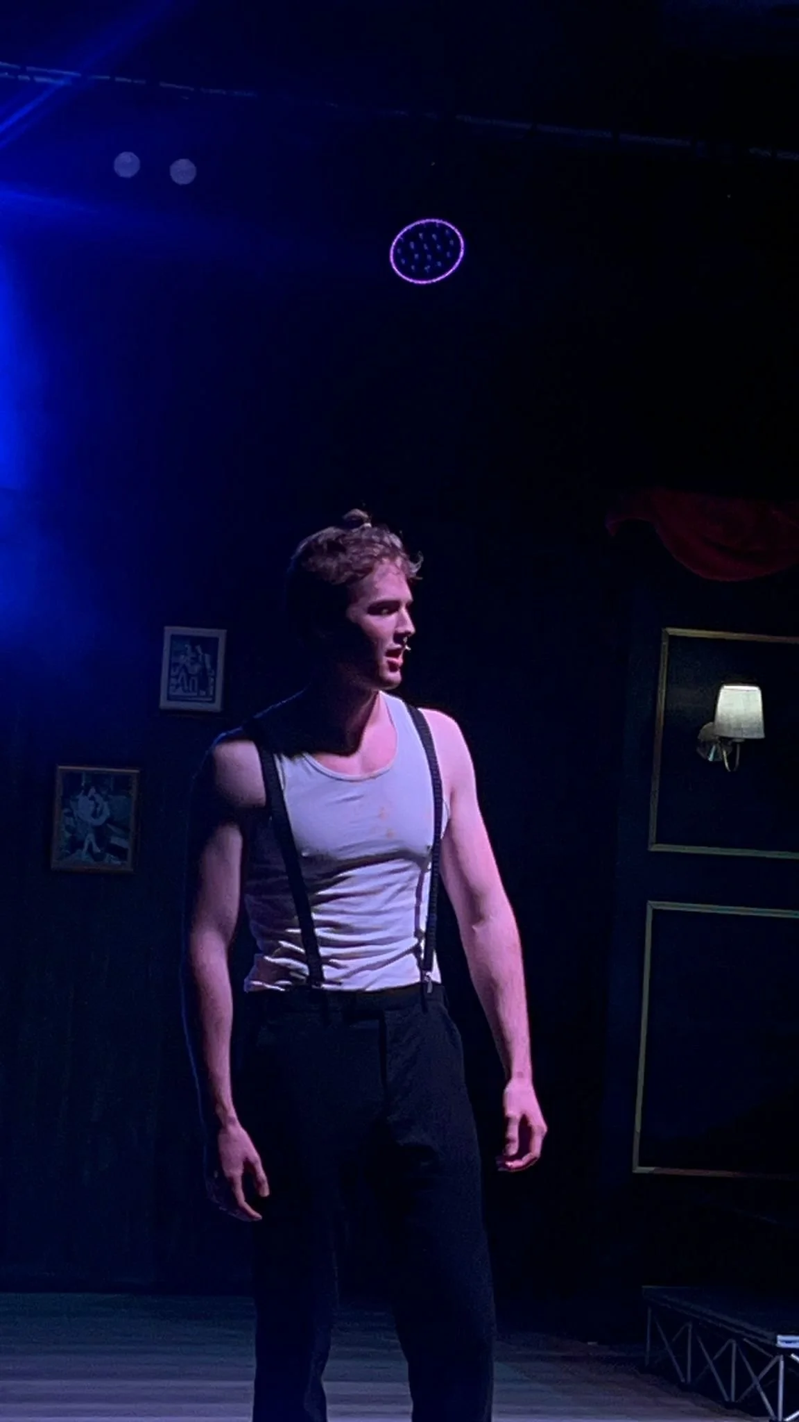 A man dressed in a white tank top with suspenders and black pants standing in a dimly lit stage or performance space.