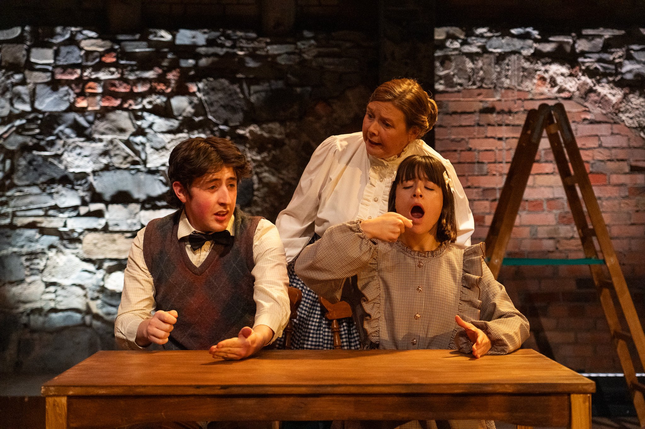 Scene from a theatrical play featuring four actors in vintage costumes, including a woman standing and talking to three seated individuals, in front of a brick and stone background.