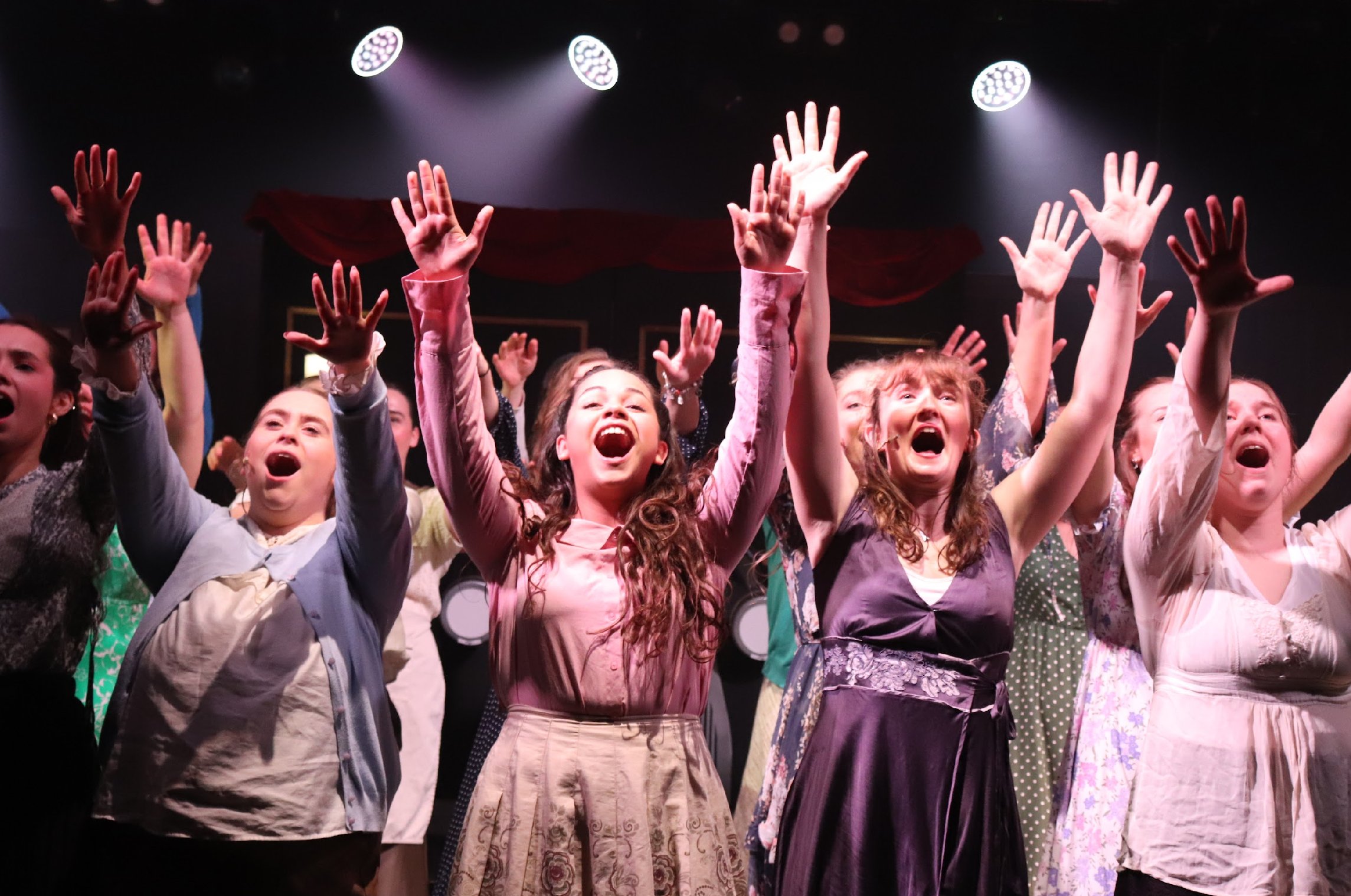 Group of young girls with raised hands in a theater or stage setting, cheering and smiling.