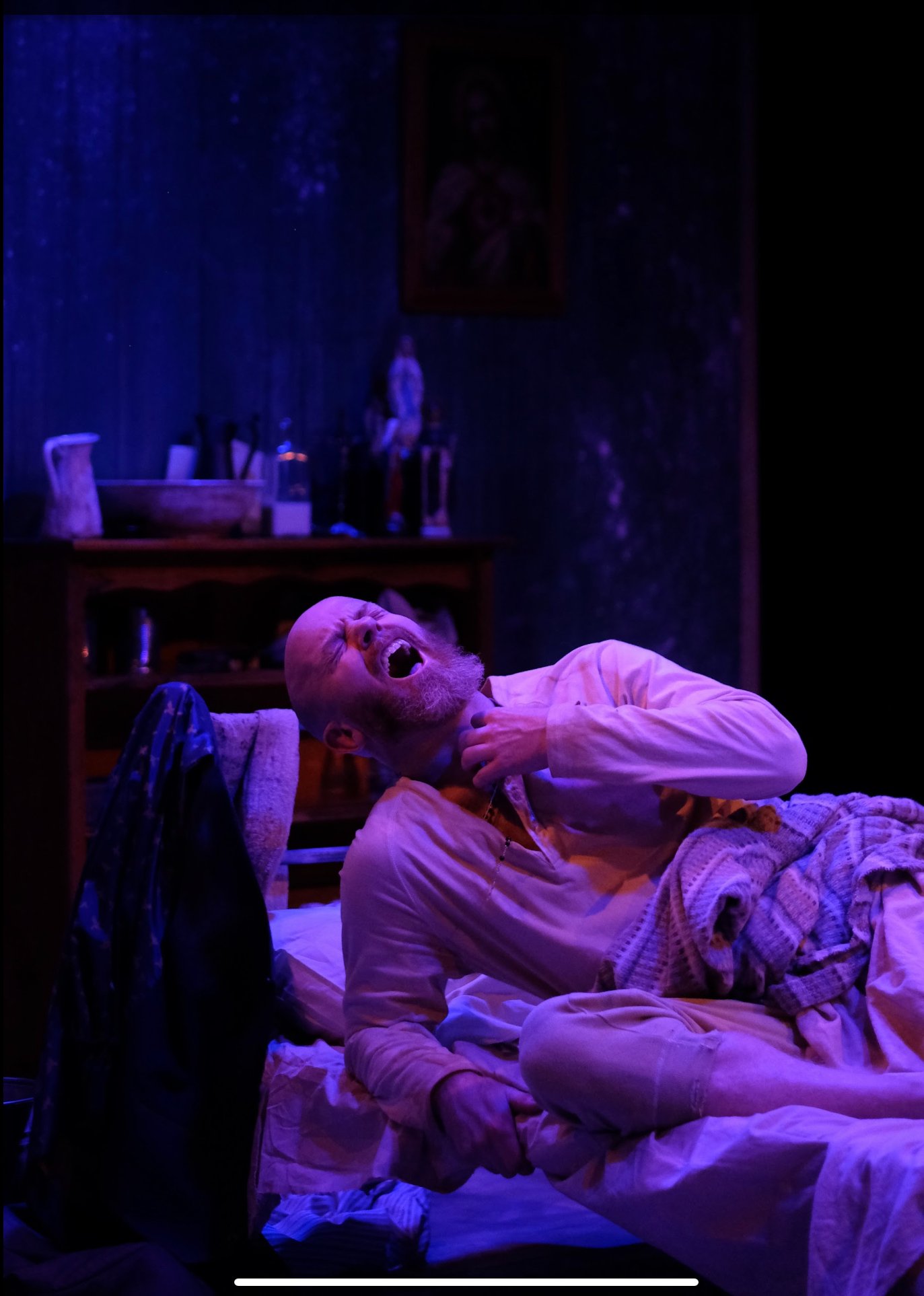 A man with a beard and light-colored pajamas lying on a bed, appearing to be in pain or distress with his mouth open and hand on his neck, in a dimly lit room with purple and blue lighting, a wooden cabinet behind him, and a framed picture hanging on