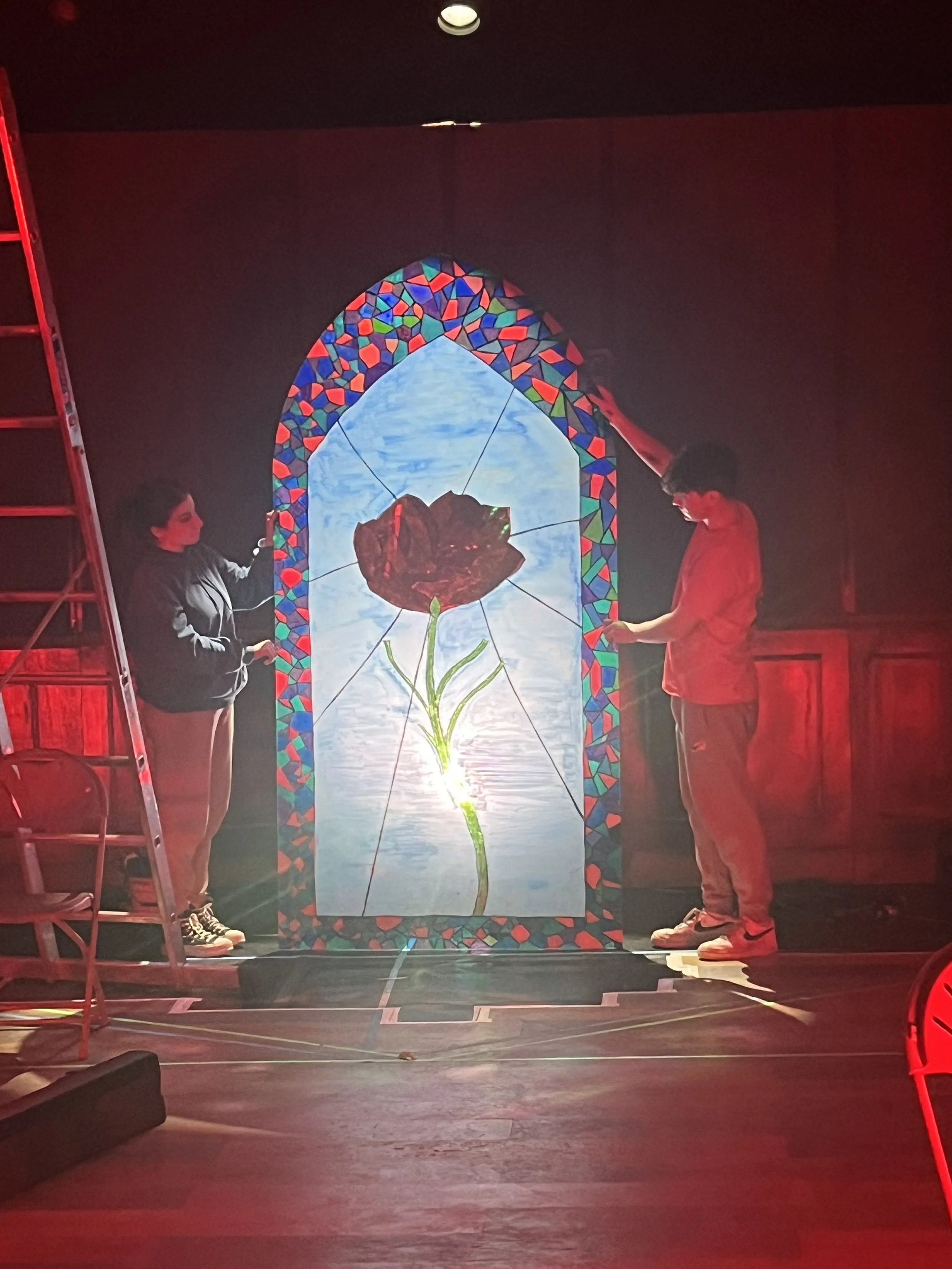 Two people standing on a stage looking at a large, pentagon-shaped stained glass window featuring a drawing of a flower. The woman on the left is wearing a gray hoodie and light-colored pants, and the man on the right is wearing a red jacket and beig
