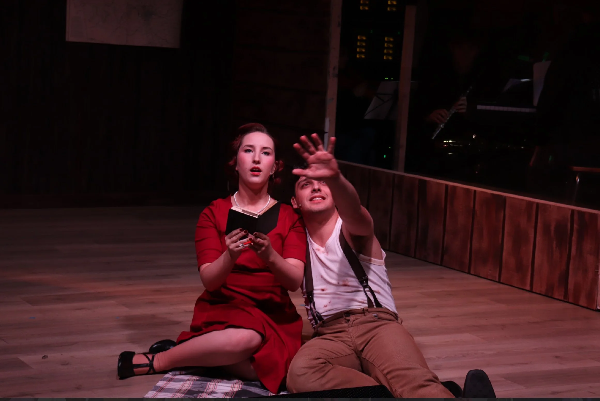 A woman with red hair in a red dress and a man with suspenders and brown pants are sitting on the wooden floor, with the woman holding a book, and the man reaching up with his hand. They appear to be in a dark indoor setting, possibly a stage or rehe