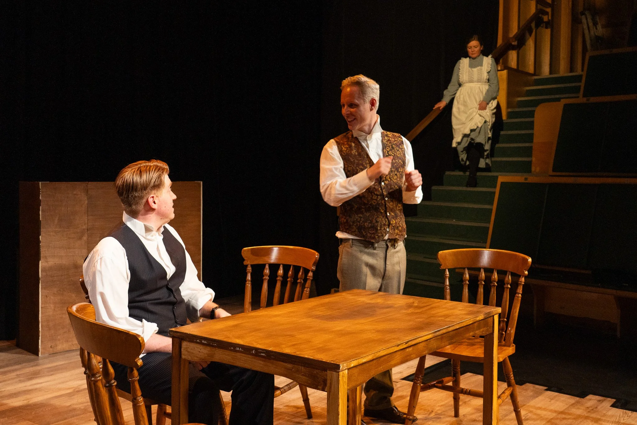 Three actors in period costumes performing a scene on stage. One man is sitting at a wooden table, another man stands nearby, and a woman is descending stairs in the background.