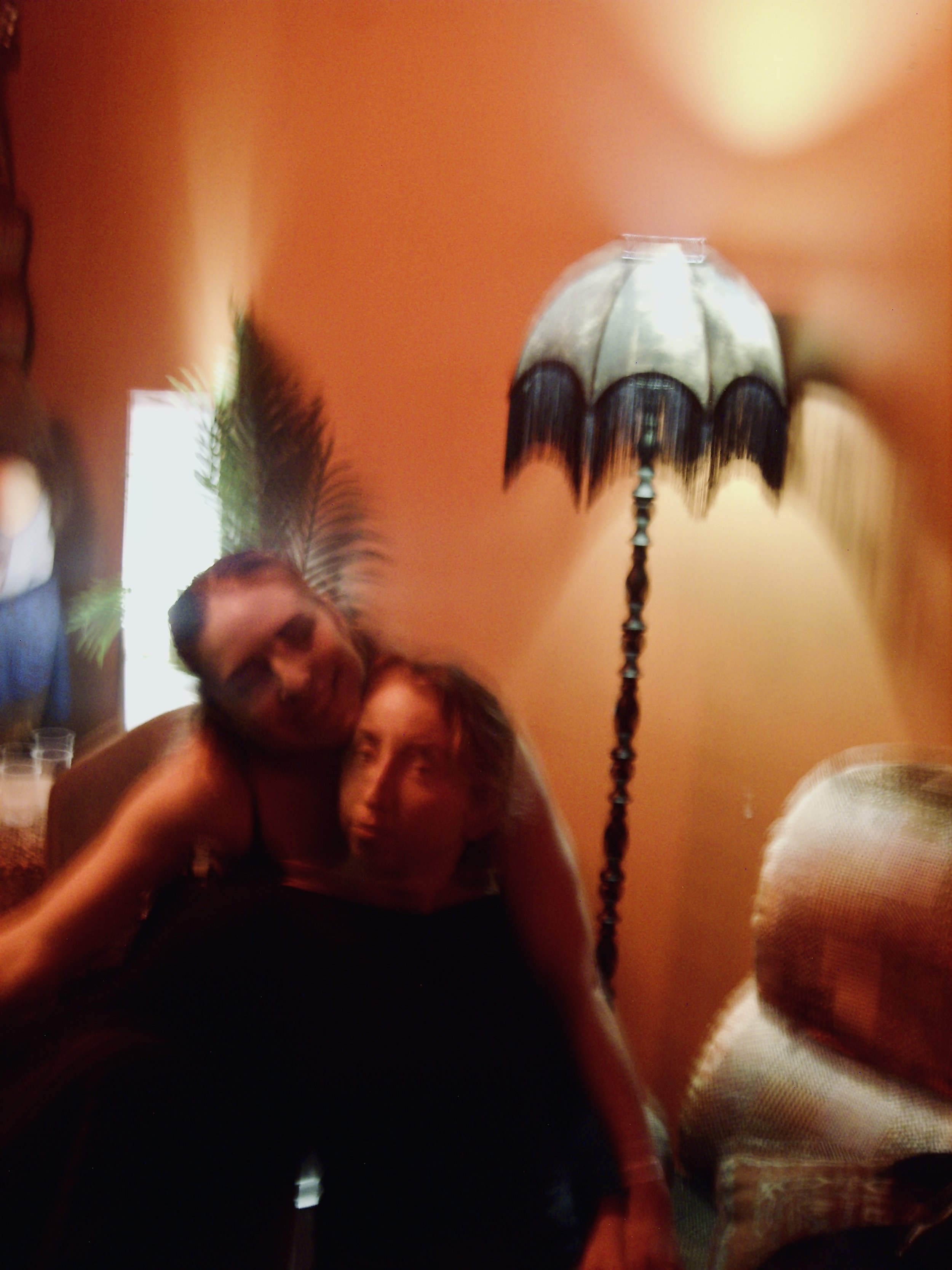A dimly lit room with peach-colored walls. In the foreground, a man with short hair and dark sunglasses is smiling and leaning close to a woman with long dark hair and a woman with short light hair. On the right side, a person with white hair is visi