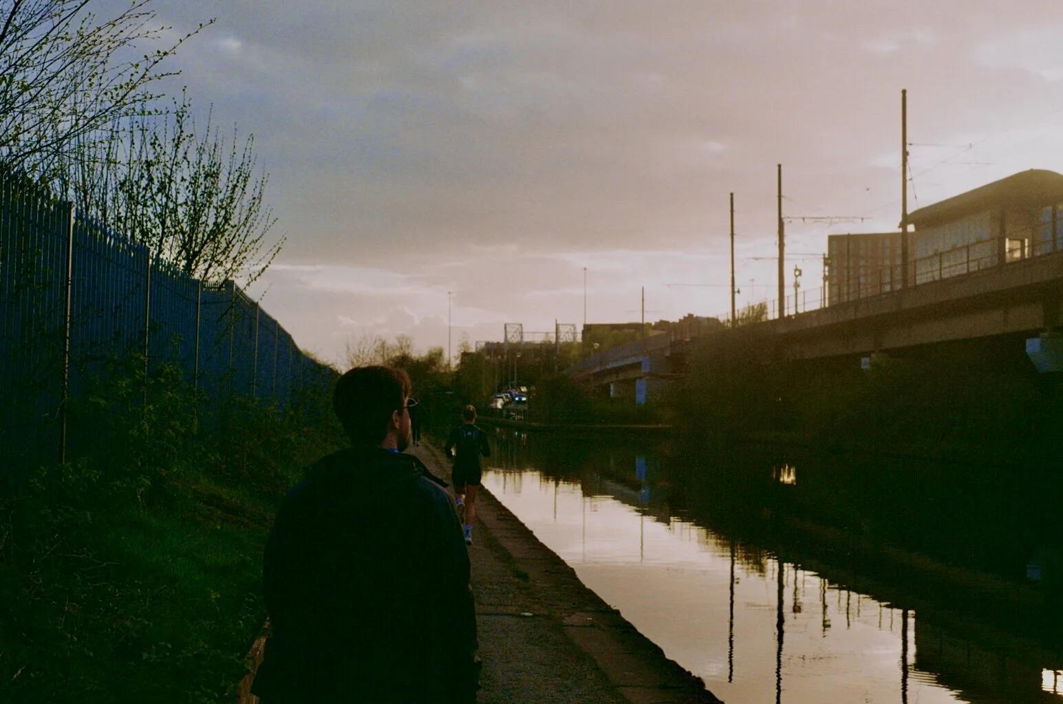 A person walking along a canal during sunset, with other runners ahead. The scene includes a bridge, train tracks, and a building in the background.