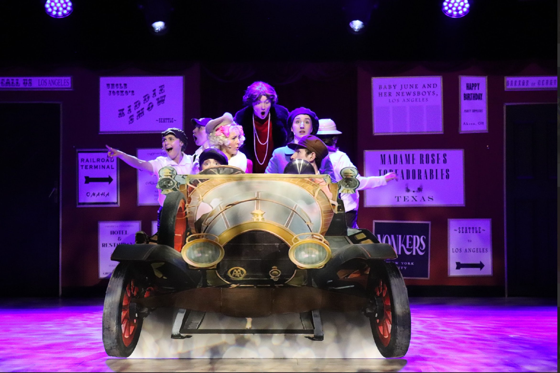 The image shows a theatrical scene with a vintage car on stage and several children in period costumes, with a woman in the background reacting with surprise or excitement. The stage background has signs with various city names and event information.