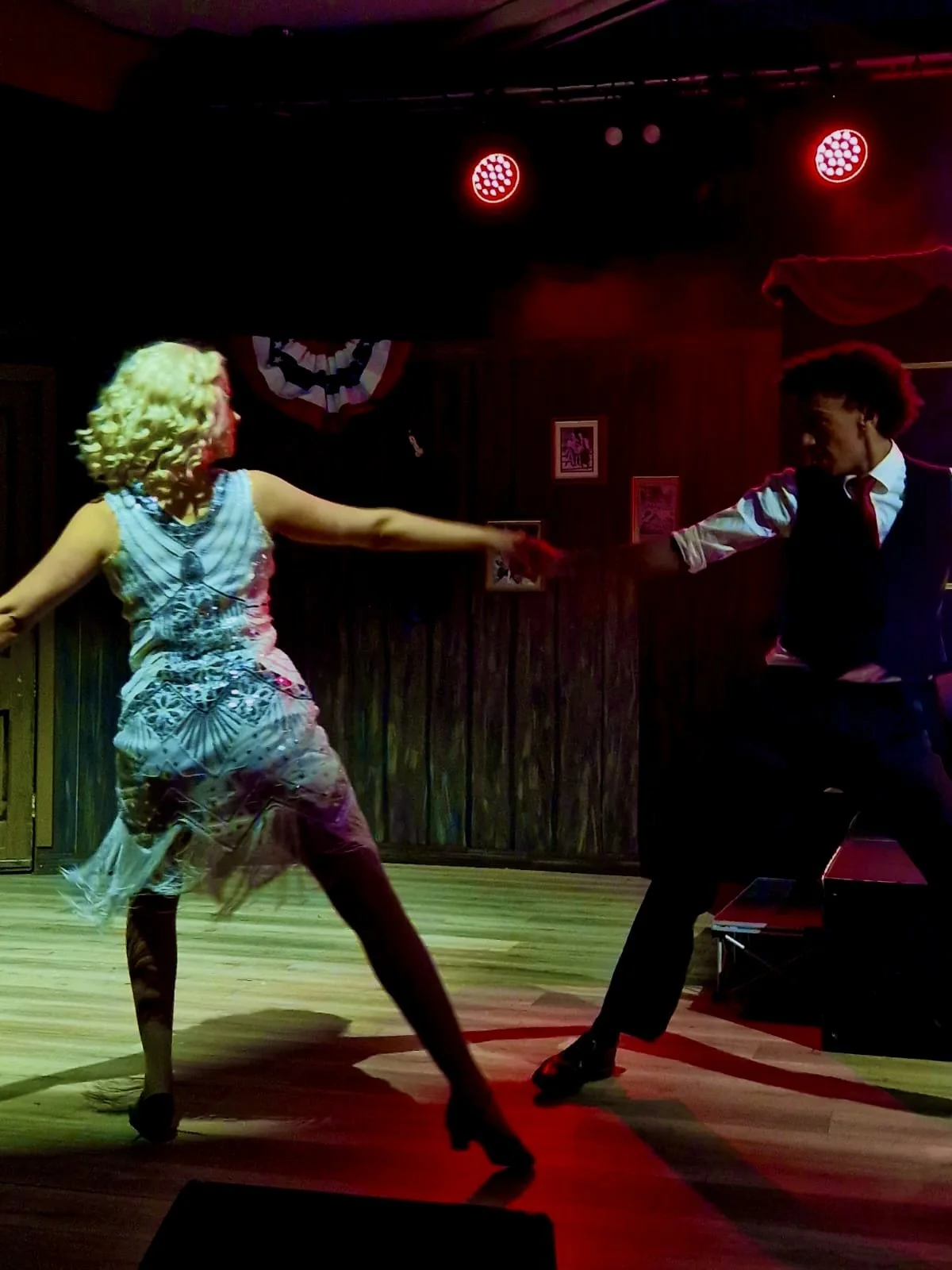 A man and woman dance together on a stage with wooden flooring. The woman is wearing a patterned dress and the man is dressed in a dark vest, white shirt, and tie. They are holding hands and appear to be performing a dance in a dimly lit setting with