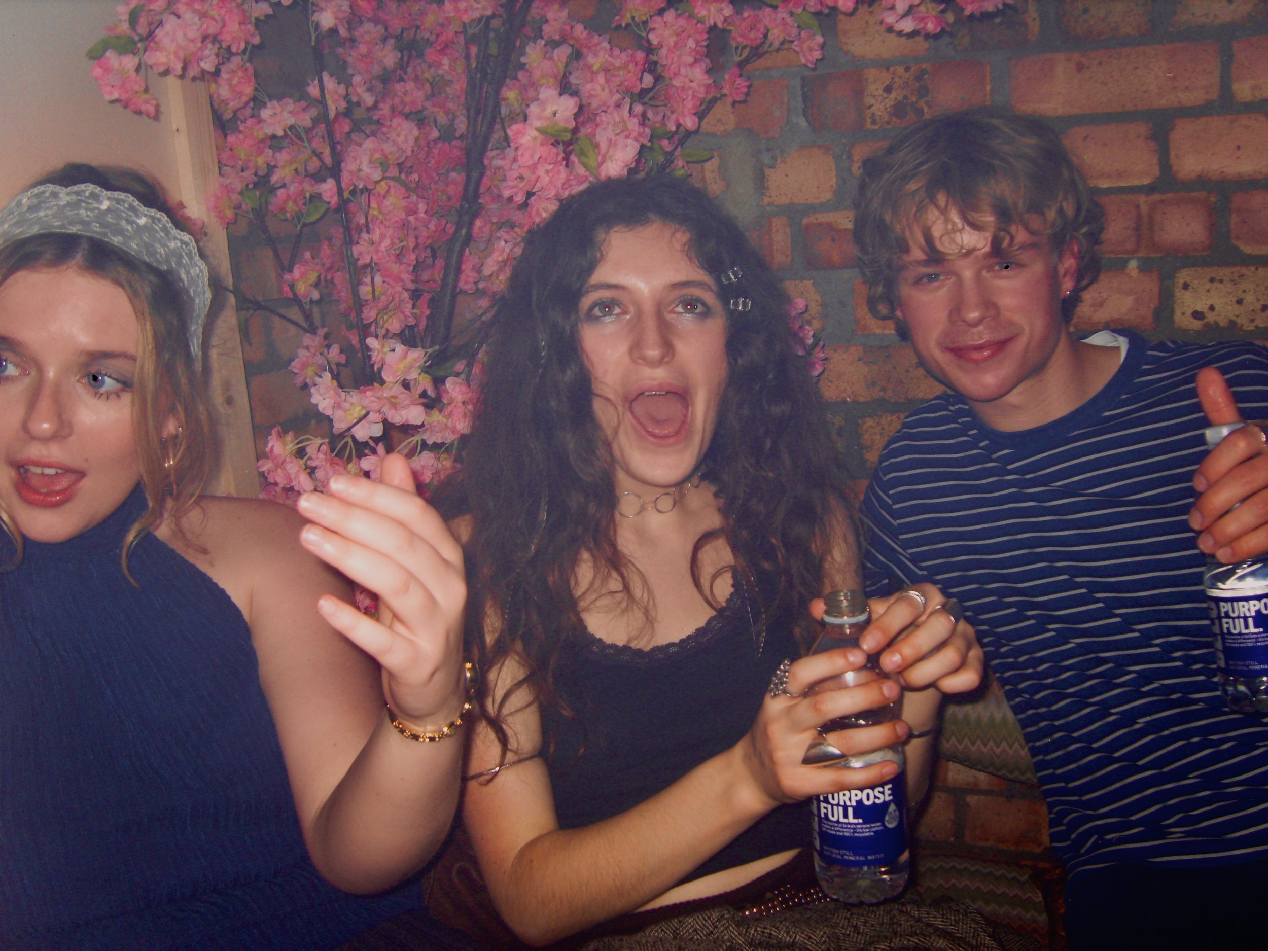 A group of three friends celebrating at a party, with a pink flowering plant and brick wall in the background. They are holding drinks, and the woman in the middle is smiling with her mouth open.