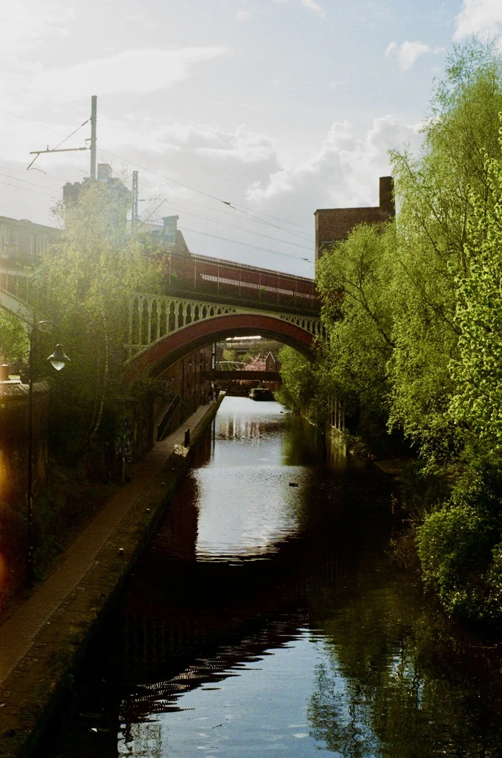 A river running under a decorative iron and brick bridge with green trees on both sides, under a blue sky with some clouds.