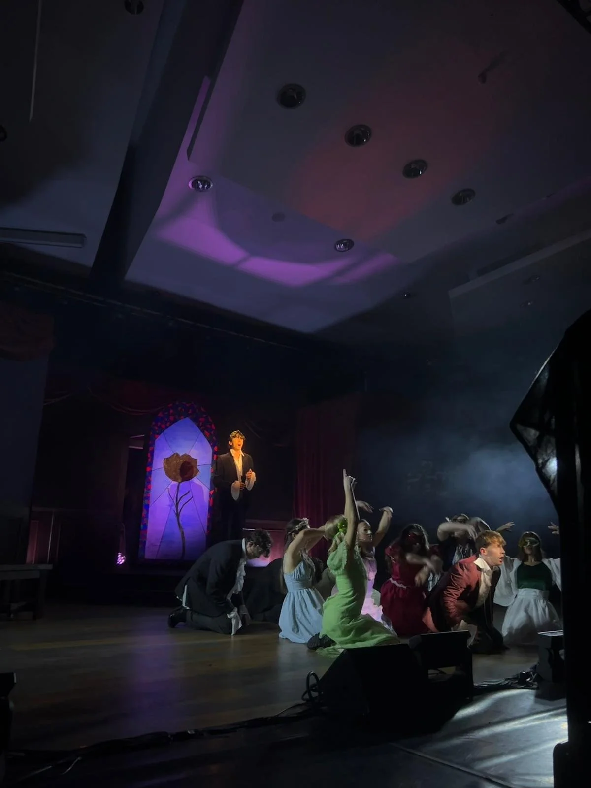 A theatrical performance featuring actors on stage, with a woman standing near a stained-glass backdrop and several performers kneeling or dancing in colorful costumes.