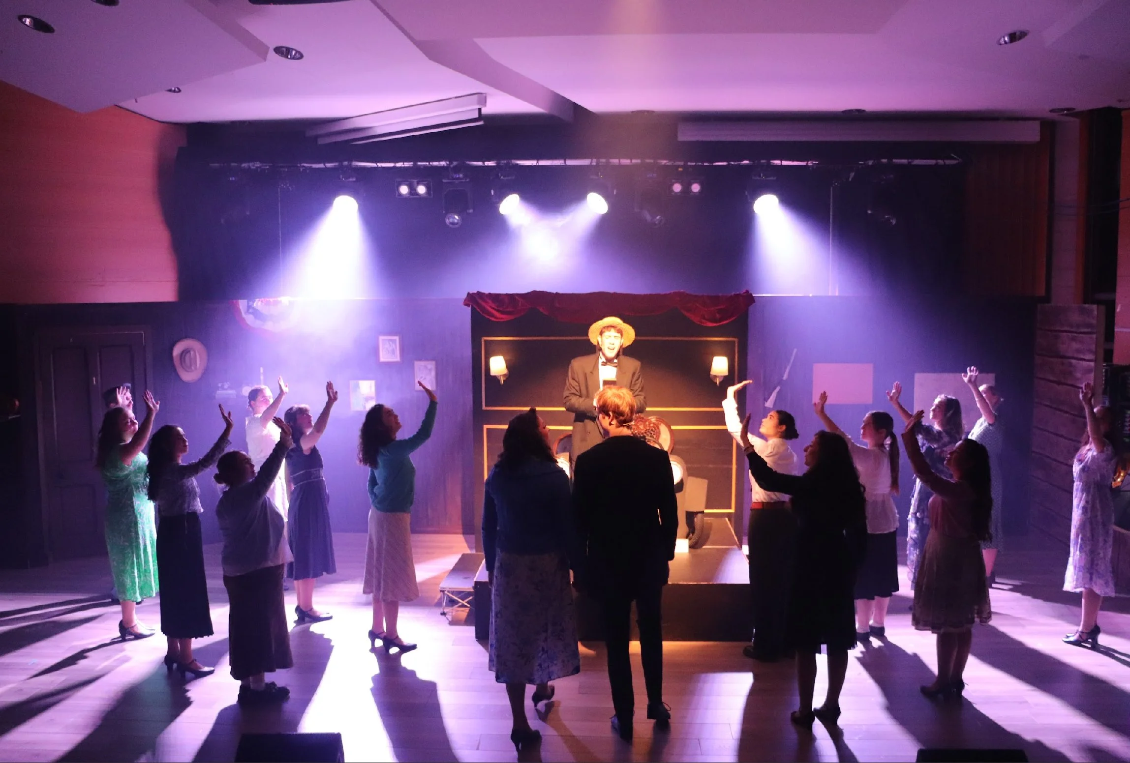Group of people dancing in a room with stage lighting, a performer in a suit and hat stands in the center, audience are raising hands and watching.