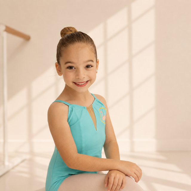 A young girl in a teal leotard sitting and smiling in a bright dance studio.