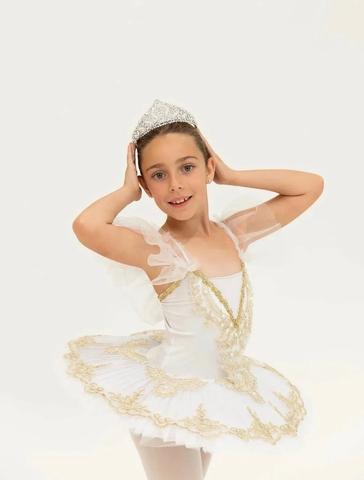 Young girl in a white ballet dress with gold details, wearing a tiara, posing with her hands near her head against a white background.