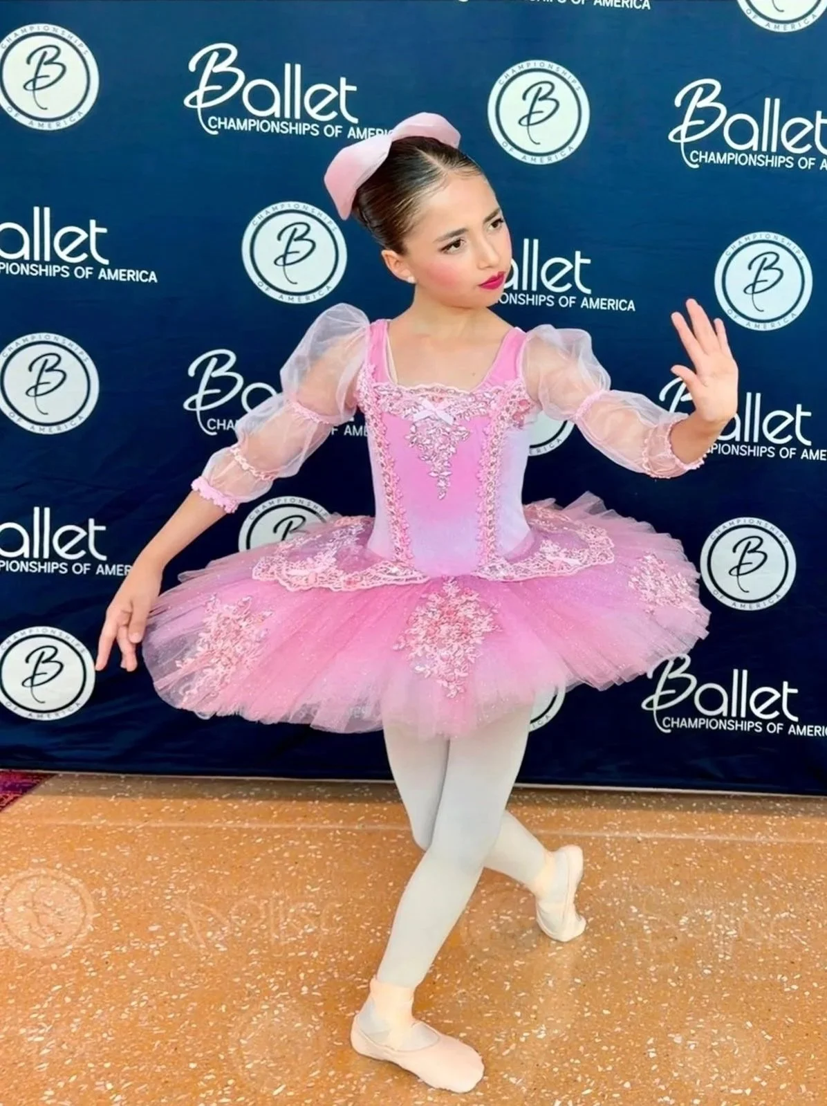 A young female ballet dancer in a pink tutu and tutu jacket, with a pink bow in her hair, rehearses or performs ballet in front of a backdrop that reads 'Ballet Championships of America'.