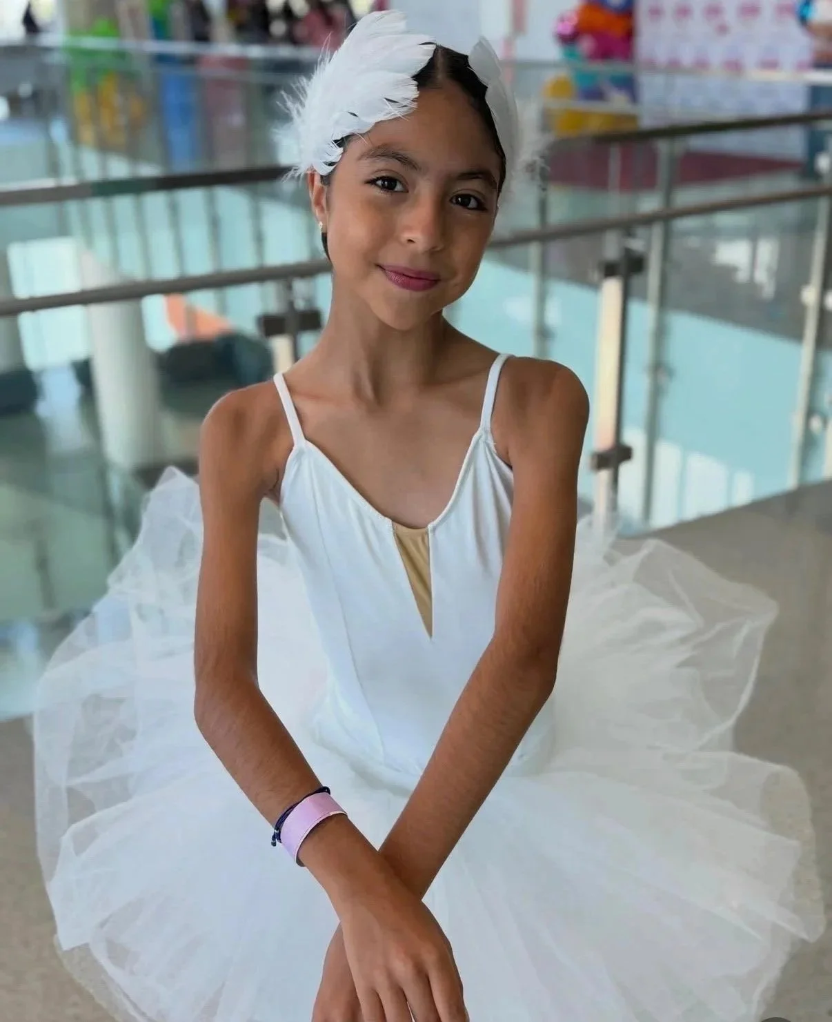 Young girl with dark hair in a braid, wearing a white dress with a tulle skirt, a feather headpiece, and a pink wristband, smiling at the camera in a brightly lit indoor setting.