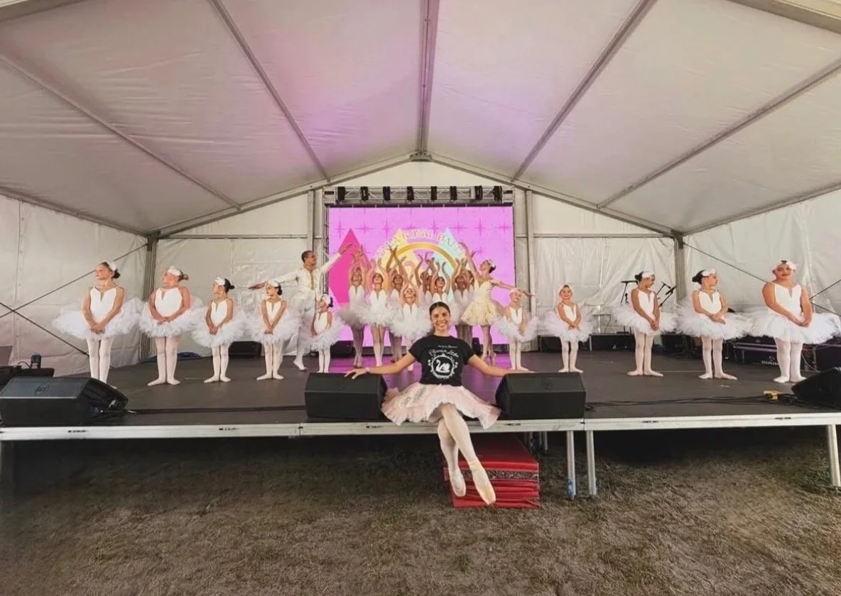 Children dressed as ballerinas on stage, posing in ballet costumes with tutus, and a woman sitting in front of the stage with a joyful expression, in a large tent.