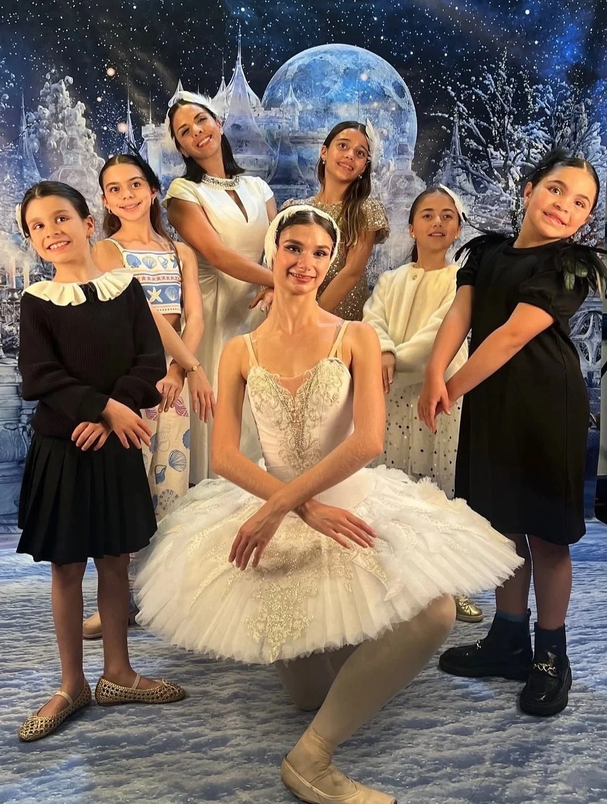 A group of seven women and girls posing in front of a winter-themed backdrop with a castle, full moon, and snow-covered trees. One woman is dressed as a ballerina, seated in front, while the others are standing around her.