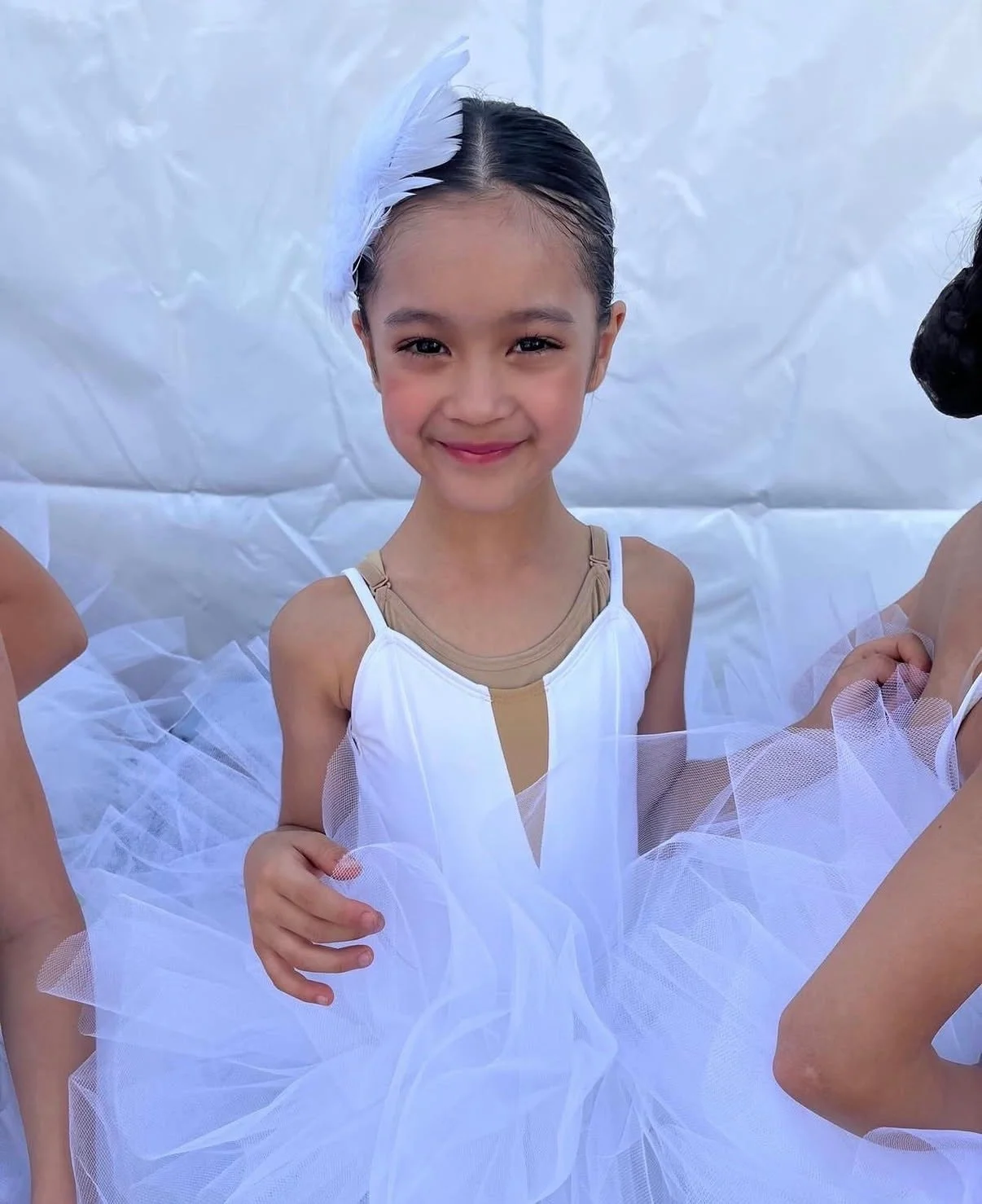 A young girl smiling in a white ballet costume with beige accents and a white feathered headpiece, standing against a light background.