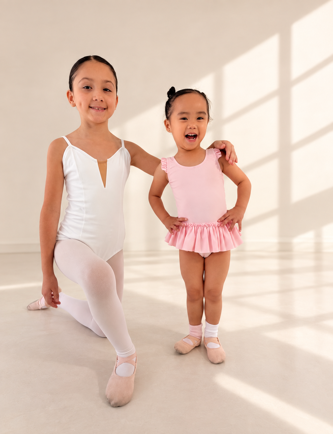 Two young girls dressed in ballet outfits standing on a light-colored floor in a room with natural sunlight and shadows on the wall behind them.