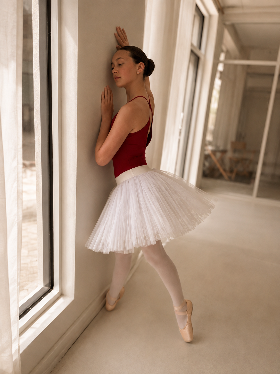 A ballet dancer in a red leotard and white tutu posing against a wall in a sunlit room, wearing pointe shoes and stockings.