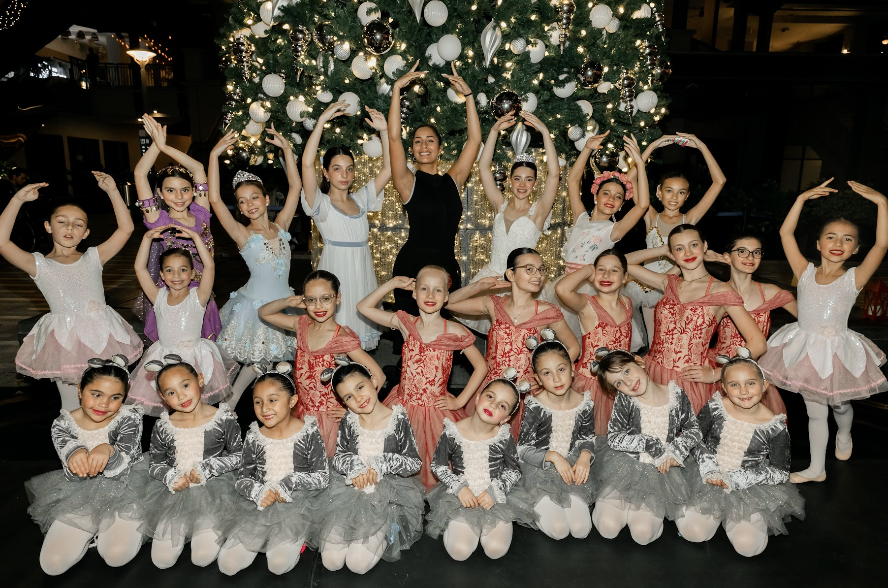 A group of young girls dressed in colorful ballet costumes and tutus, posing on stage in front of a large decorated Christmas tree with white and silver ornaments and lights.