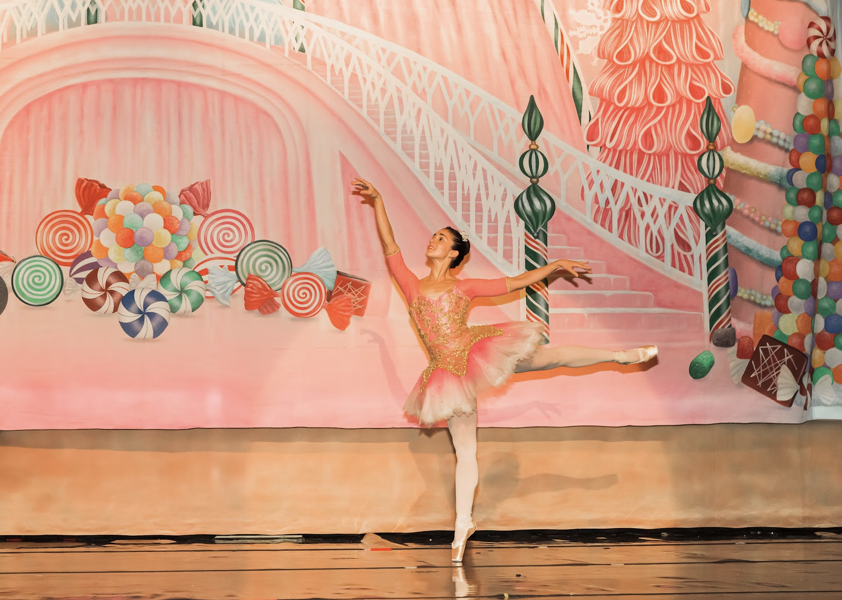 A ballet dancer in a pink and gold tutu performing on stage with a colorful candy-themed backdrop.