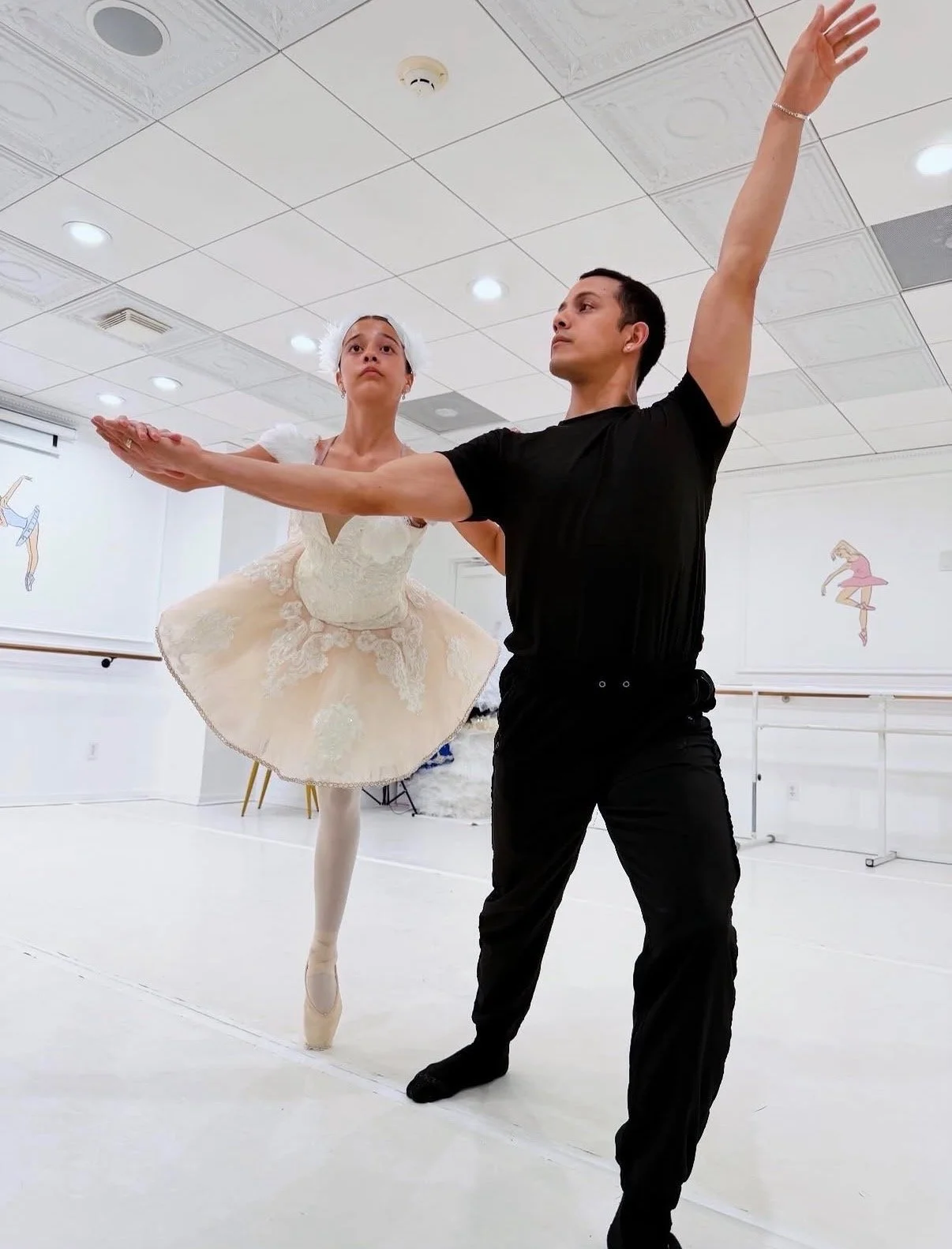 A male ballet instructor practicing with a female ballet dancer who is on pointe, wearing a tutu, in a dance studio with ballet-themed decorations on the wall.