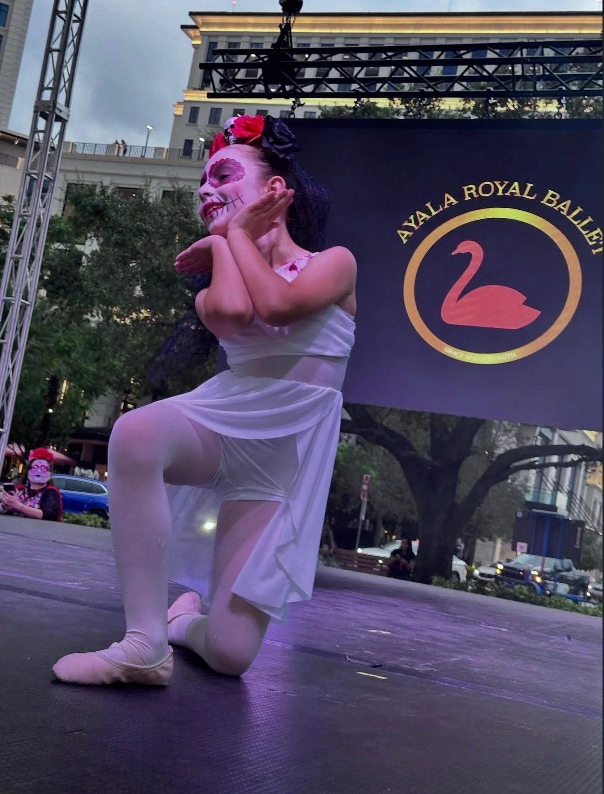 Performer dressed as Frida Kahlo with sugar skull makeup, in a white dress, dancing on stage at an event for Azalea Royal Bali, with a logo featuring a pink swan on a dark background.