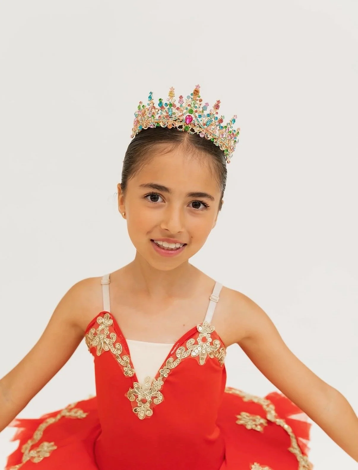 Young girl dressed in a red ballet costume with gold embellishments, wearing a colorful jeweled crown, smiling, against a plain white background.