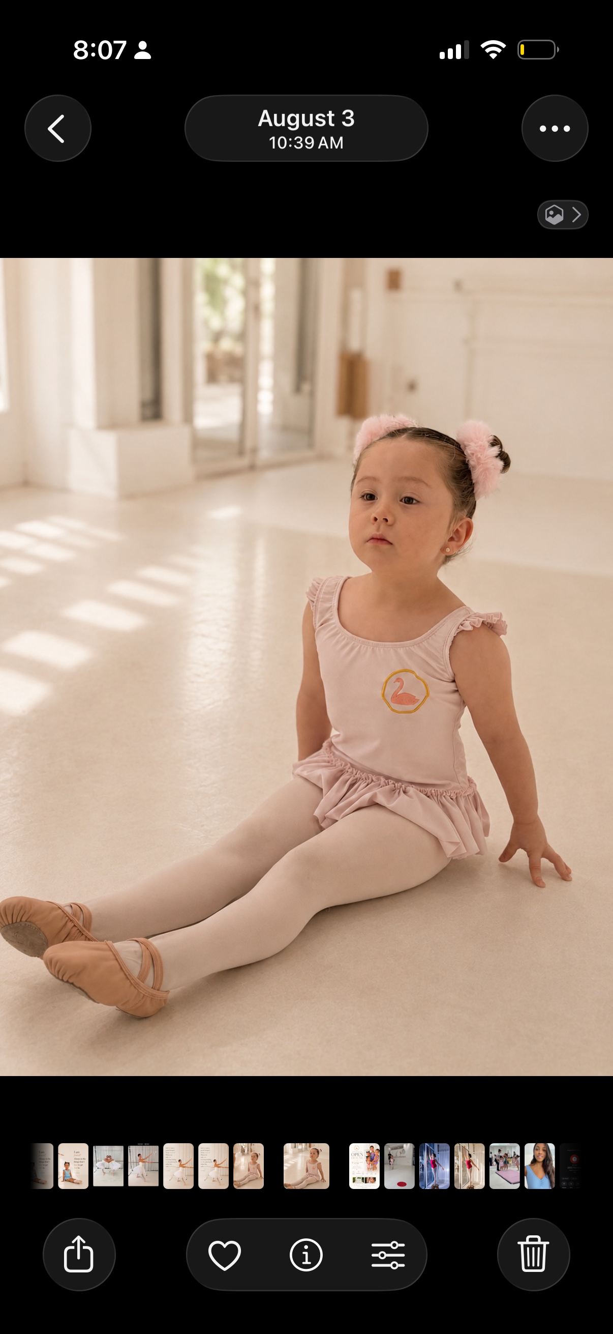 A young girl sitting on the floor in a ballet studio, wearing a pink ballet outfit with a swan emblem, pink ballet slippers, and pink hair accessories, with light streaming through windows behind her.