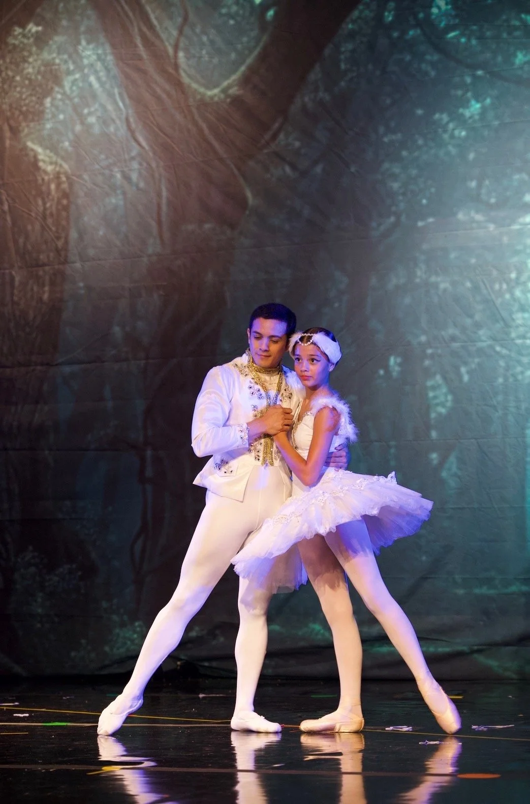 Two ballet dancers, a young man and young woman, performing on stage. They are dressed in elegant white costumes with gold accents for the man and a white tutu for the woman, with a white headpiece. They are holding hands and standing close together,