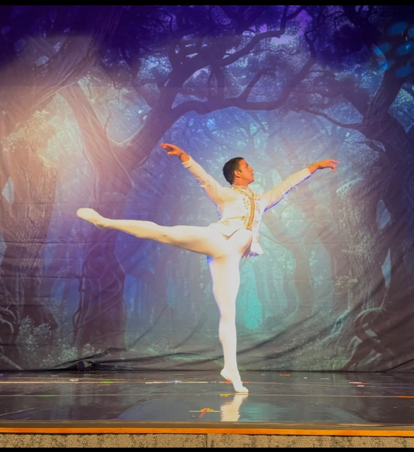 A male ballet dancer in a white costume performing a balance pose on stage with a forest backdrop.