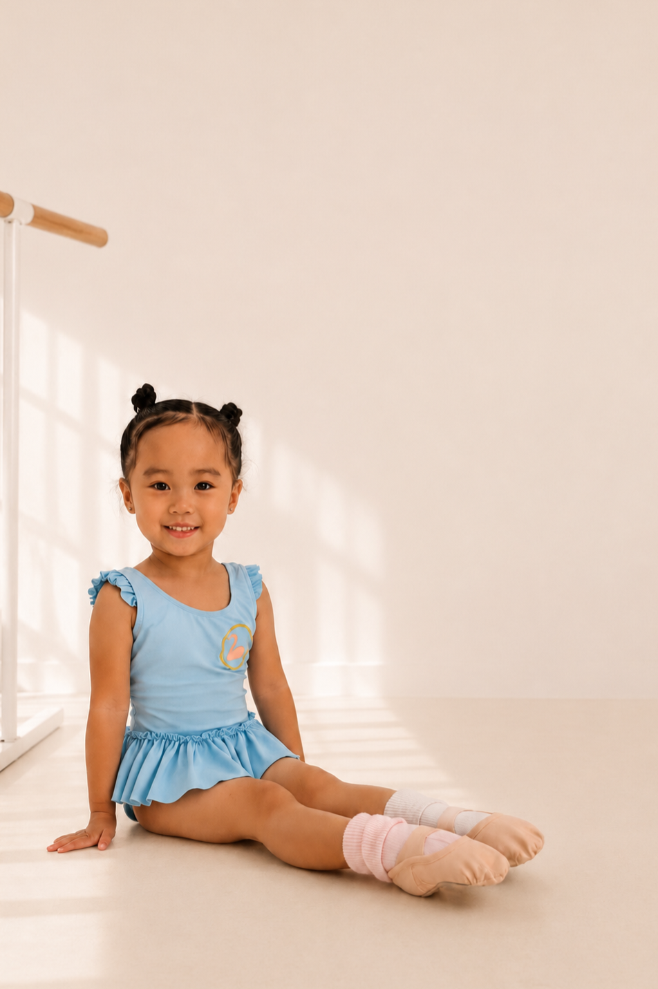 Young girl sitting on a dance studio floor, smiling, wearing a light blue dance outfit, ballet slippers, and knee-high pink and white socks.