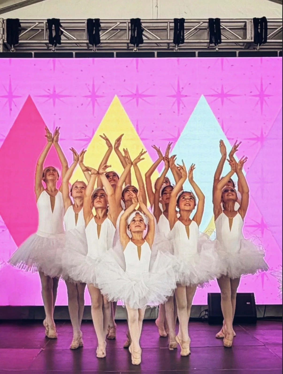 A group of young ballerinas in white tutus performing on stage with colorful geometric background and purple lighting.