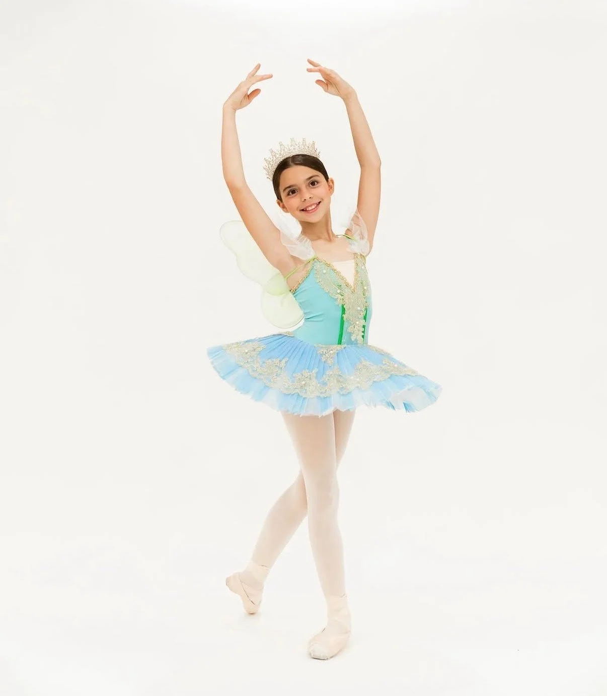 Young girl dressed as a ballerina fairy in a colorful tutu and fairy wings, posing with arms raised.