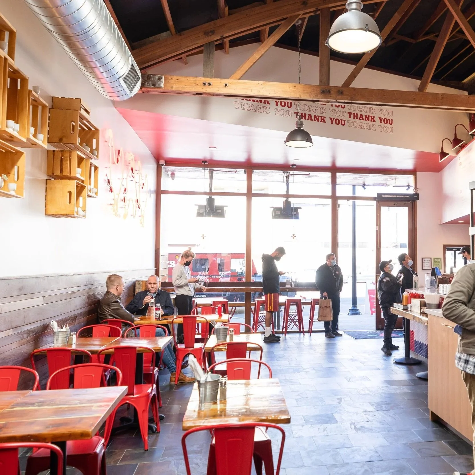 Inside Proposition Chicken in San Francisco with red chairs and wooden tables. Customers are dining and standing in line at the counter for order. Large windows let in natural light, and a wall displays a 'Thank You' message. 