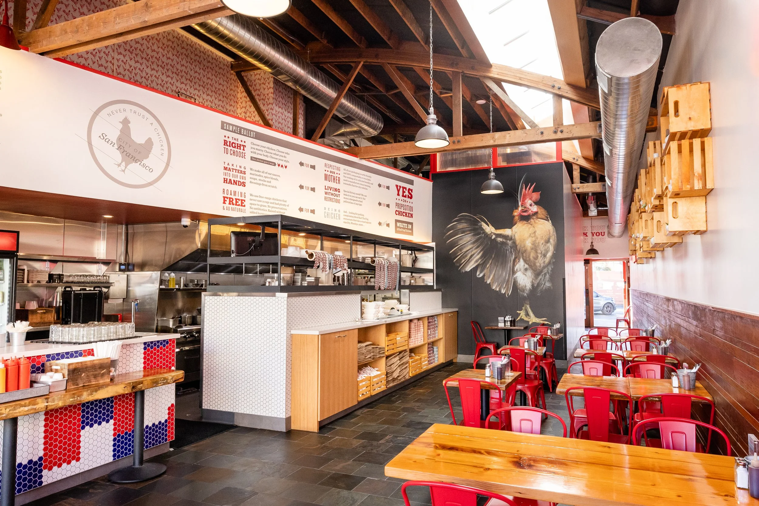 Indoor view of Proposition Chicken in San Francisco with wooden beams, ductwork, and red chairs; a menu board with chicken options and a large cartoon rooster illustration on the wall.