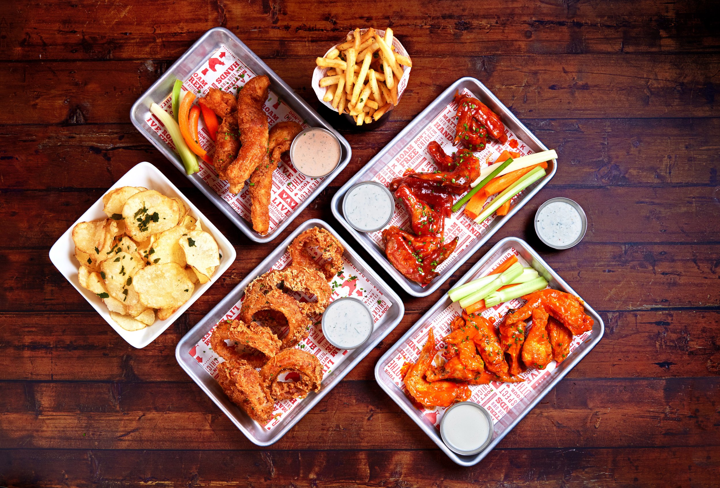 Assorted fried foods including chicken wings, onion rings, and potato chips served with dipping sauces on metal trays and a bowl of French fries on a wooden table.