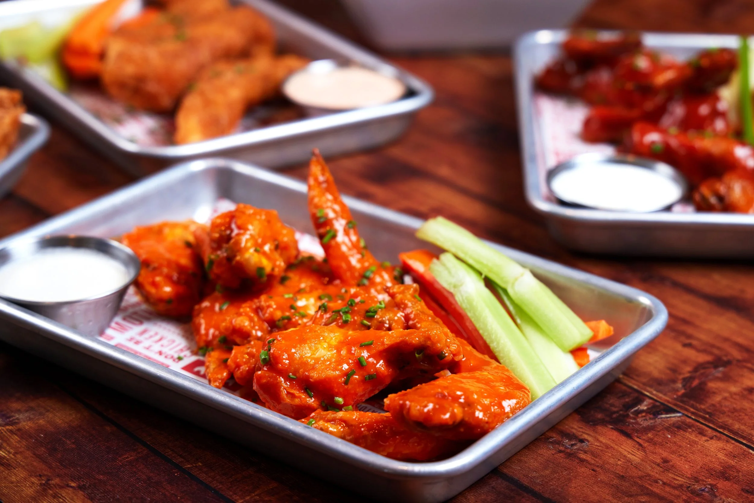 Tray of buffalo chicken wings garnished with chopped chives, served with celery sticks and carrot sticks, with dipping sauces on the side.